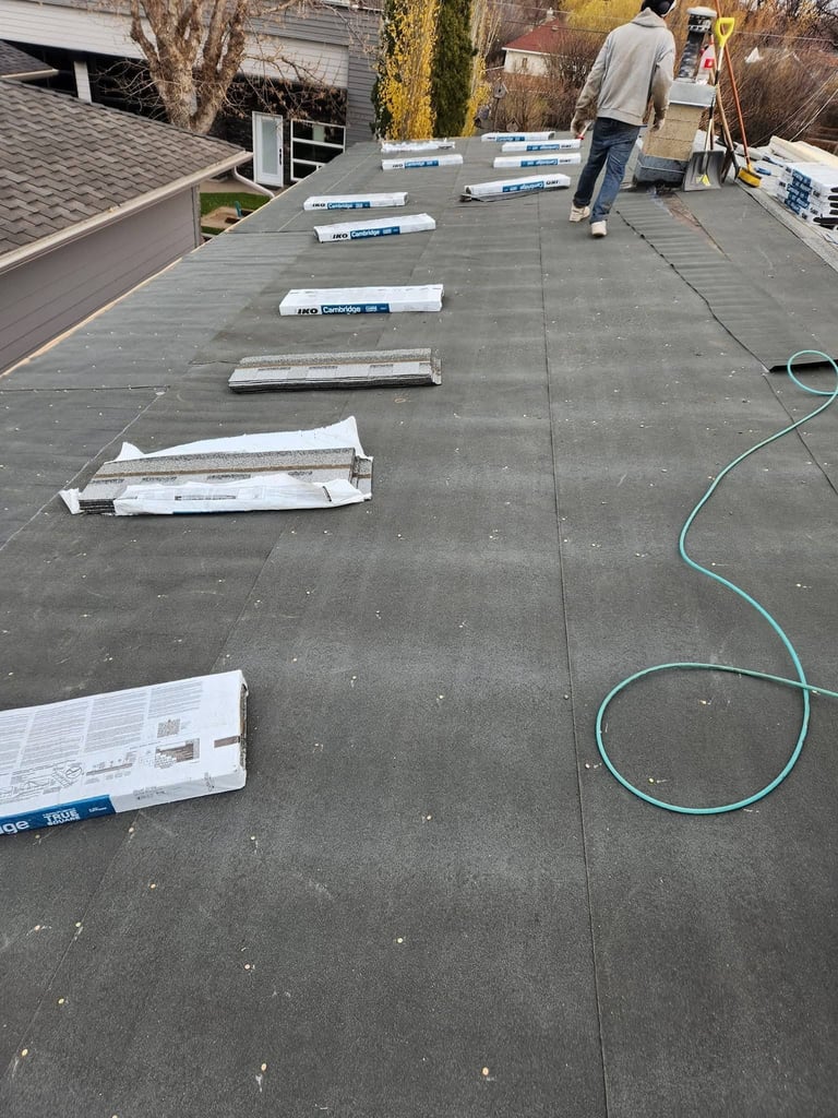 Roofing materials and supplies laid out on a flat roof during installation, with a worker visible in the background and a green hose on the ground