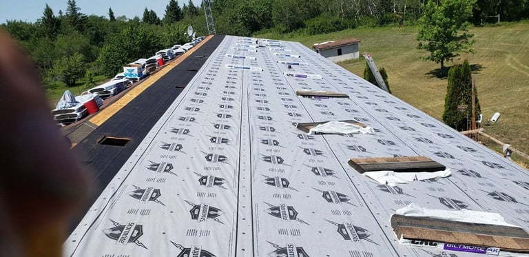 Roof being installed with protective underlayment membrane and wooden framing visible, residential construction site with vehicles and trees