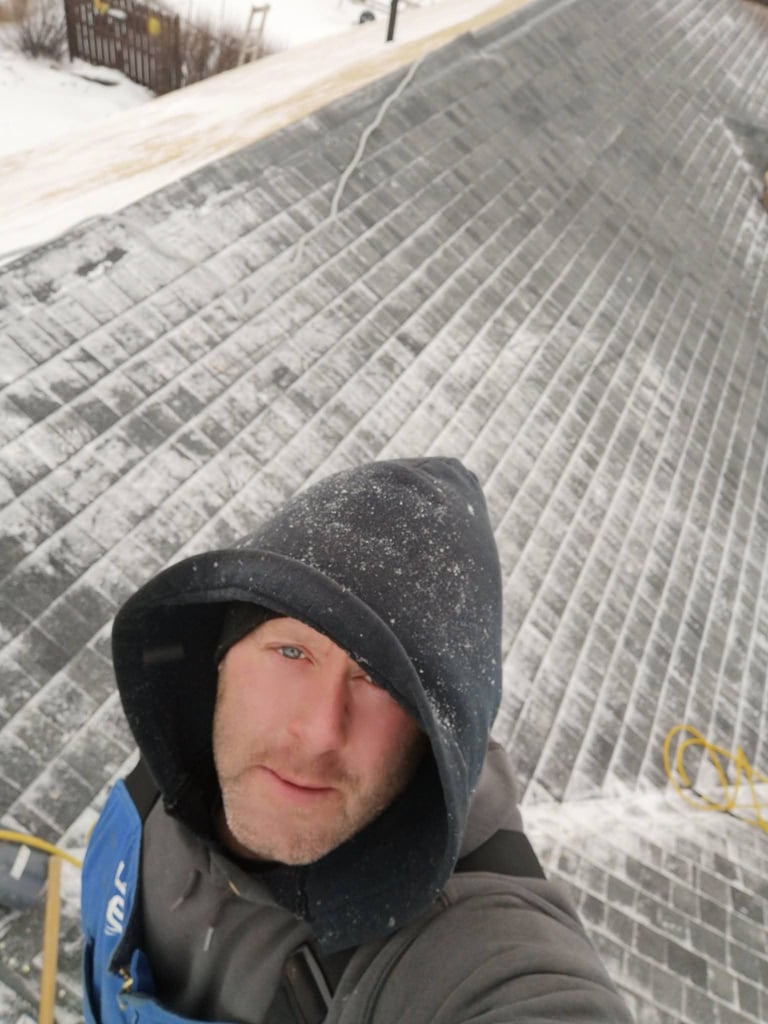 Man wearing black beanie and dark jacket smiles at camera while standing on snowy roof with metal grating pattern