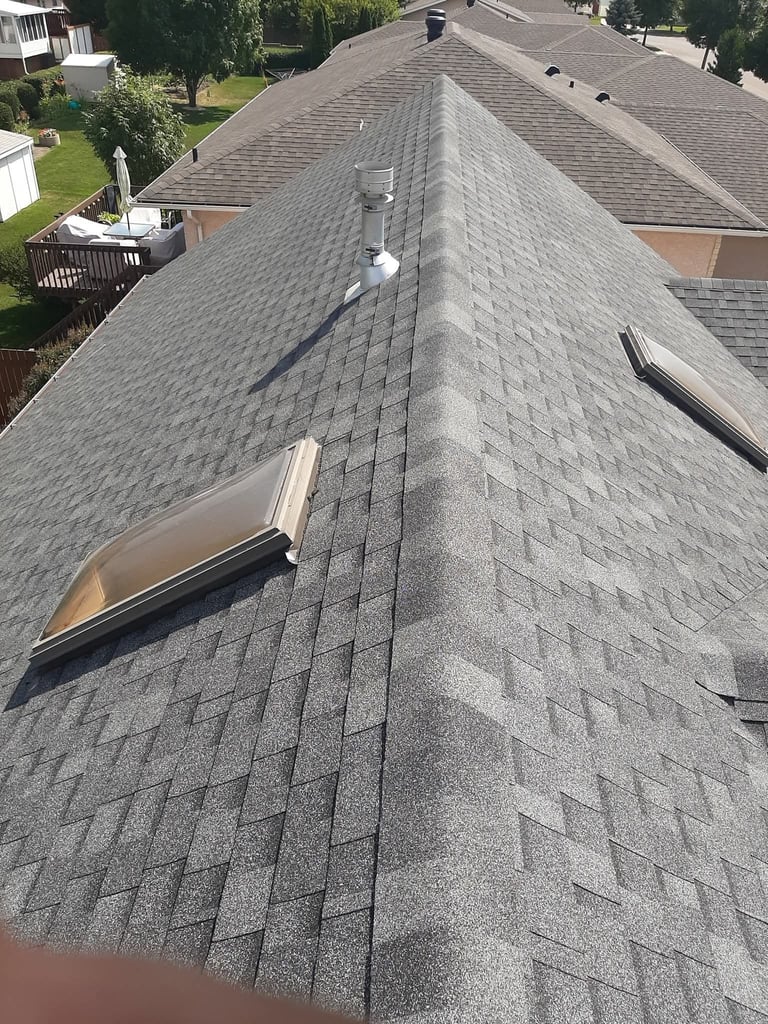 Aerial view of a residential roof with gray asphalt shingles, a skylight, and vent pipes in a suburban neighborhood
