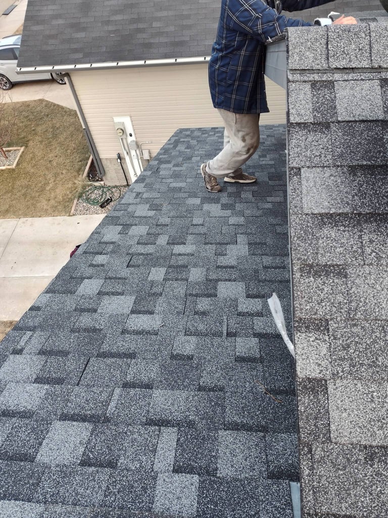 Person in blue plaid shorts standing on gray asphalt shingles during roof installation or inspection work